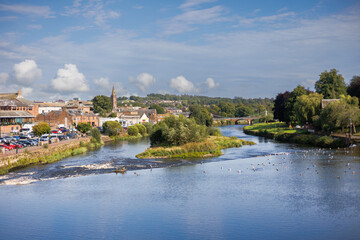 Obraz premium Scenic view of a river flowing through Dumfries, Scotland, taken from a bridge, with reflections and surrounding urban and natural landscape
