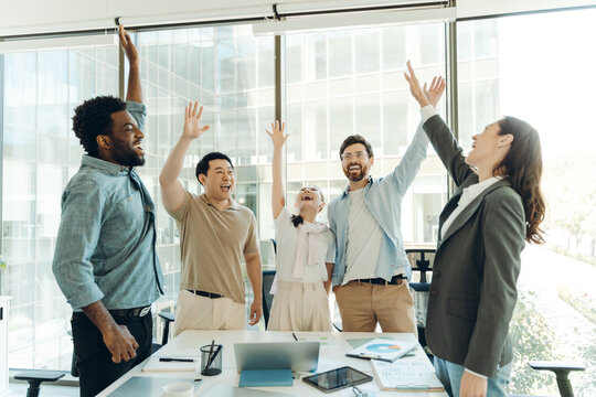 Diverse group of happy business team celebrating success raising hands in office