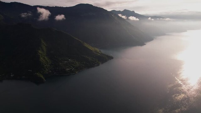 A breathtaking high-altitude drone shot captures a long pier extending into the glassy, reflective waters of Lake Atitl&aacute;n at sunrise.