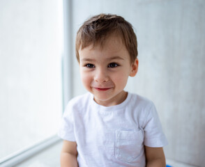 Naklejka premium Smiling boy in casual clothing indoors. A young boy with short brown hair smiles happily in a bright indoor setting, wearing a simple white shirt.