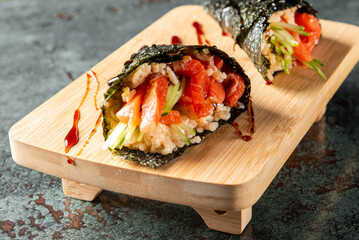 Two hand-rolled sushi cones with raw fish, rice, cucumber, and nori on a wooden board, garnished with sauce. Japanese cuisine, fresh and artistic food presentation