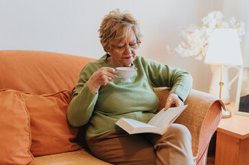 A serene elderly woman reading a book while sipping tea, relaxing on a comfortable sofa in a warmly lit room with soft orange decor and a cozy ambiance.