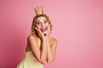 Adorable young woman in a yellow dress posing with a crown on a pink background, expressing joy and charm
