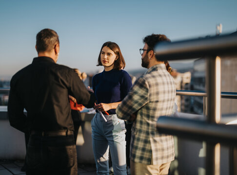 Business colleagues engage in a brainstorming session on a rooftop balcony at sunset, embracing diversity and creativity in a relaxed atmosphere. The multicultural team discusses innovative ideas