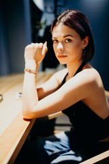 Young woman with short dark hair sitting at a table, looking into the camera with a thoughtful expression. Portrait of a stylish girl in casual outfit, illuminated by warm light indoors.