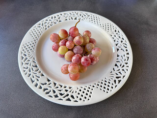 Decorative white carved plate holding a bunch of pink grapes on a dark surface, photographed from above, elegant and minimal still life composition.