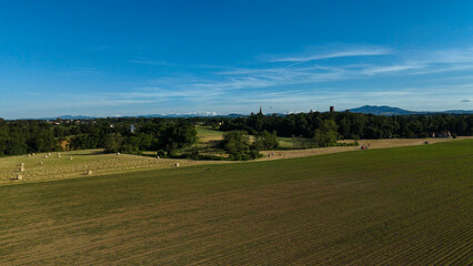 Fototapeta premium Aerial view capturing farmer collecting hay bales via tractor near Revigliasco, Italy, with Monviso and Alpine peaks towering against bright blue summer sky
