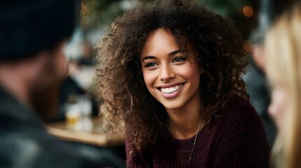A happy young woman with curly hair smiles brightly while engaging in a lively conversation with friends at a casual cafe setting