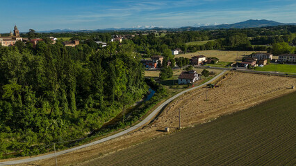 Aerial panorama revealing verdant Italian landscape, stone village nestled among patchwork farmlands, serpentine road, Monviso mountain rising against azure summer sky