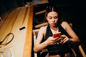 A young woman sits at a wooden table in a dimly lit room, focused on her phone. Dressed in a black top and jeans, she shows a calm yet concentrated mood, illuminated by warm light.