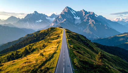 An aerial view of a long, winding road traversing a lush green mountain ridge, leading towards majestic snow-capped peaks under a clear sky.