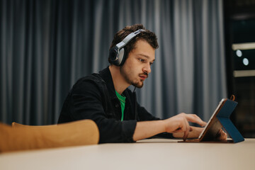 A teenage boy wearing headphones is deeply focused on using a tablet. The indoor setting and relaxed posture suggest a casual or learning-related activity with a contemporary atmosphere.