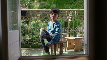 Young boy sitting on a wooden stool on a balcony, smiling and holding his foot, surrounded by...