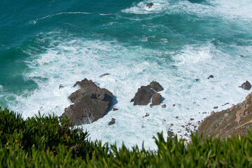 Foaming ocean waves are crashing against the rocky coastline of Cabo da Roca, Portugal. The westernmost point of continental europe, vibrant green vegetation in the foreground. Wallpaper background