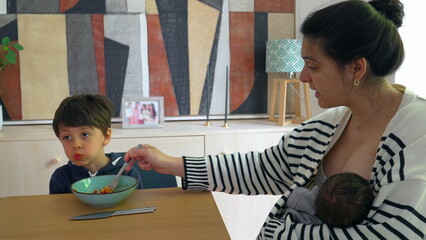 Woman feeding spaghetti to her son while breastfeeding newborn in a cozy home environment, illustrating the busy life of a mother caring for multiple children simultaneously