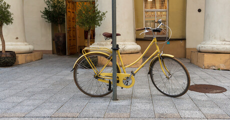 Fototapeta premium Classic yellow bicycle with brown seat and metal frame locked to a pole in front of a historic building with columns. The retro design and muted colors add charm to an urban street scene.