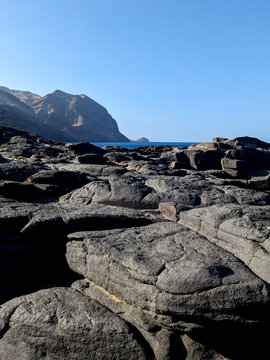 Dramatic Coastal Landscape with Weathered Volcanic Black Rocks, Rugged Mountain Range, and Blue Ocean under Clear Sky