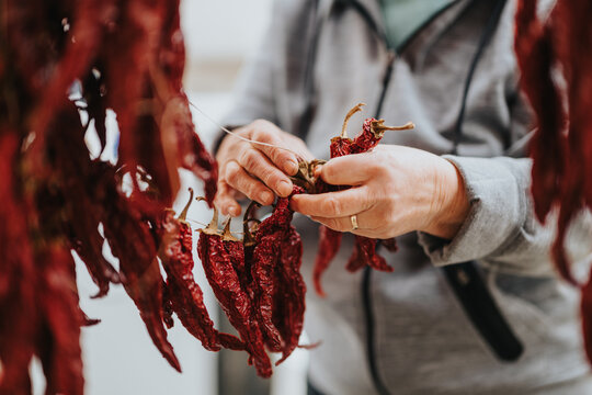 Close-up of an elderly woman's hands threading vibrant dried red chili peppers on a string, highlighting cultural and culinary traditions in food preparation.