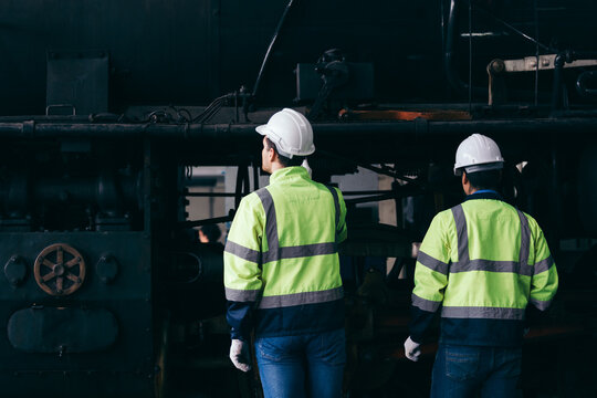 Two railway engineers wearing reflective safety jackets and helmets inspect an old locomotive engine in maintenance facility, emphasizing teamwork, inspection routine and industrial operations. - Powered by Adobe
