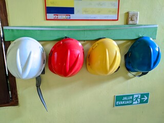 Colorful safety helmets hanging on a wall, including white, red, yellow, and blue hard hats, symbolizing workplace safety and protection. Seen near an evacuation route sign indoors.