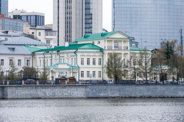 Historic government building with green roof on embankment of the Iset River in Yekaterinburg, contrasted with modern glass skyscrapers in the background