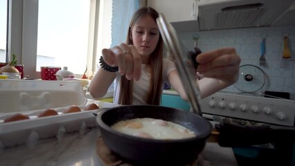 Teenage girl breaking raw egg into frying pan in home kitchen, preparing scrambled eggs for breakfast. Concept of cooking, healthy lifestyle, child independence. - Powered by Adobe