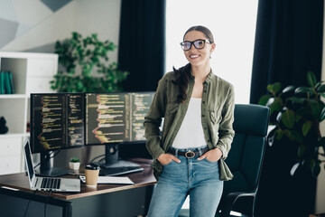 Charming professional female programmer working in a home office, standing confidently near a coding setup and workspace