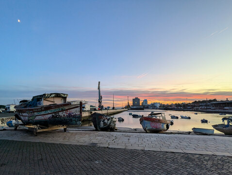 Urban Harbor Sunset with Abandoned Graffiti Boats, Calm Water, City Skyline, and Industrial Crane