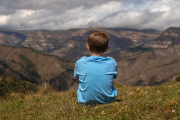 Naklejka premium Rear view of a boy sitting on a rock with mountains in the background