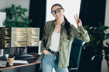 Young female programmer smiling and flashing a peace sign in her modern office workspace with coding screens