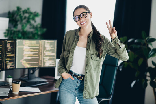 Young female programmer smiling and flashing a peace sign in her modern office workspace with coding screens - Powered by Adobe