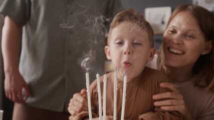 Mother removing her hands from younger sons face, he blowing out five candles on birthday cake, overjoyed family clapping