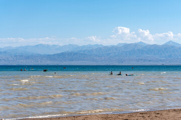 People swim and to paddleboard and boats on a mountain lake on a hot sunny summer day near a sandy beach.