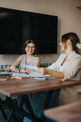 Women are sitting at a table, sharing a moment of collaboration in an office setting. They appear to be enjoying their conversation while reviewing documents, highlighting teamwork and communication.
