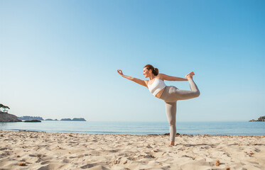 Calm woman standing on one leg doing Natarajasana or Dancer Pose and meditating in early morning hours on sandy beach with calm sea waves. Mental health, people relaxing, traveling concept