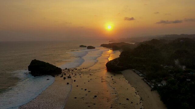 Aerial sunset over Watu Karung Beach in Pacitan, Indonesia, captures the golden hour light reflecting on the wet sand. Waves crash against rock formations, with the serene shoreline visible