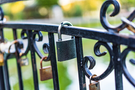 Locks attached to a fence at a park. Several locks dangle from a decorative fence near a calm pond in a public park during daylight.