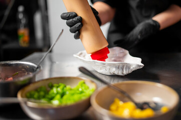 Chef wearing black gloves prepares food in a professional kitchen, dispensing sauce into a dish with fresh chopped vegetables in bowls. Culinary process, close-up