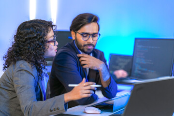 A senior software developer or team lead points at code on a screen, mentoring a female colleague. Diverse professionals collaborating on a project in a modern IT office.