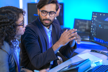 A senior software developer or team lead points at code on a screen, mentoring a female colleague. Diverse professionals collaborating on a project in a modern IT office.