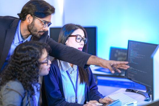 A senior software developer or team lead points at code on a screen, mentoring a female colleague. Diverse professionals collaborating on a project in a modern IT office.