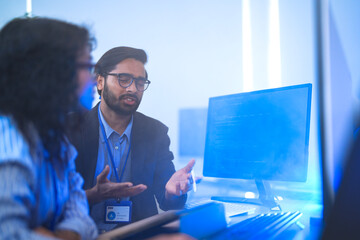 Diverse software developers in a lively discussion about a project. A male and female colleague brainstorm ideas and debate strategy in a modern, blue-lit tech office.
