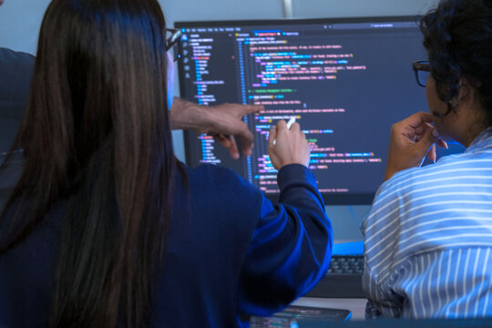 A diverse team of office workers collaborates on a project. A manager points at the computer screen, discussing the work with his colleagues in a modern office at night.