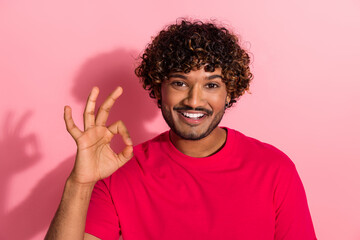 Happy young man in a red t-shirt gestures okay and smiles against a pink background with a cheerful expression