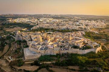Fototapeta premium An aerial view of Mdina, the historic fortified city in Malta, surrounded by ancient stone walls and medieval architecture. The golden evening light enhances the beauty of the old city.