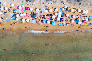 An overhead drone view shows a lively summer beach filled with colorful umbrellas, sunbathers, and swimmers enjoying the clear water. 