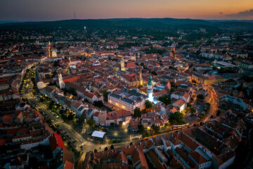 An aerial view of a historic city at dusk, illuminated by warm streetlights and surrounded by rolling hills. The old town’s narrow streets, church towers, and red rooftops create a nice landscape.