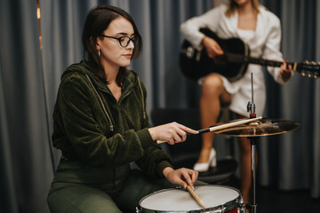 Two female musicians collaborating in a rehearsal studio, one playing drums and the other playing...