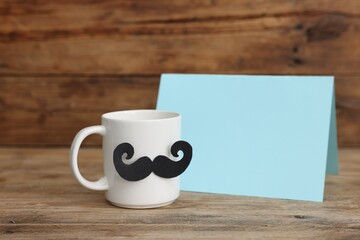 Happy Father's Day. Cup with mustache and card on wooden table. Space for text