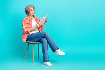 Mature Woman in Checkered Shirt Sitting on Chair and Reading a Book Against a Bright Turquoise Background